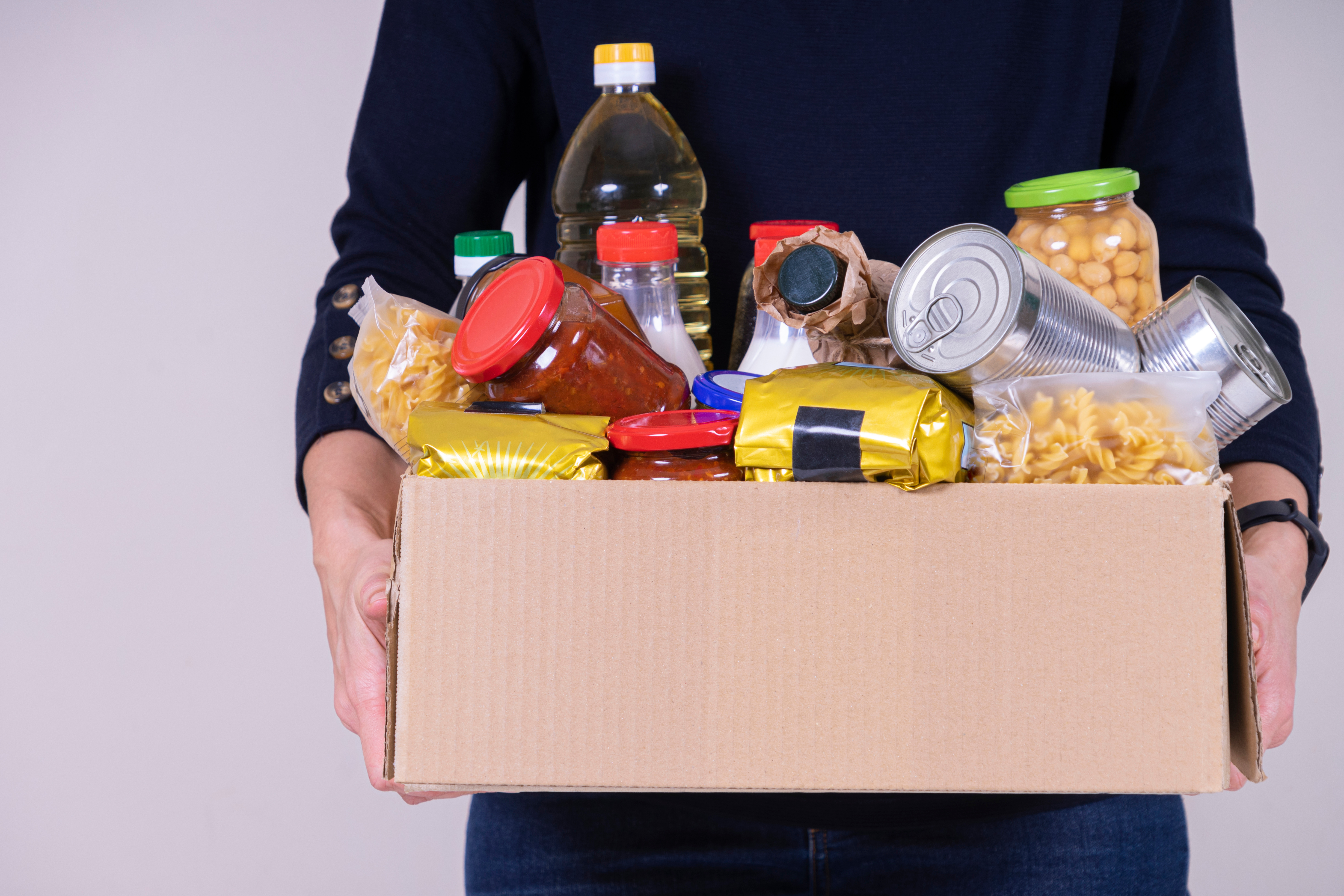 Person holding a box of donated food items including tins, pasta and jars